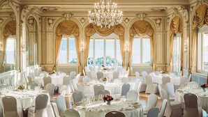 Un salón de baile amplio con candelabros ornamentados, ventanas grandes con cortinas y mesas redondas dispuestas para un evento formal.