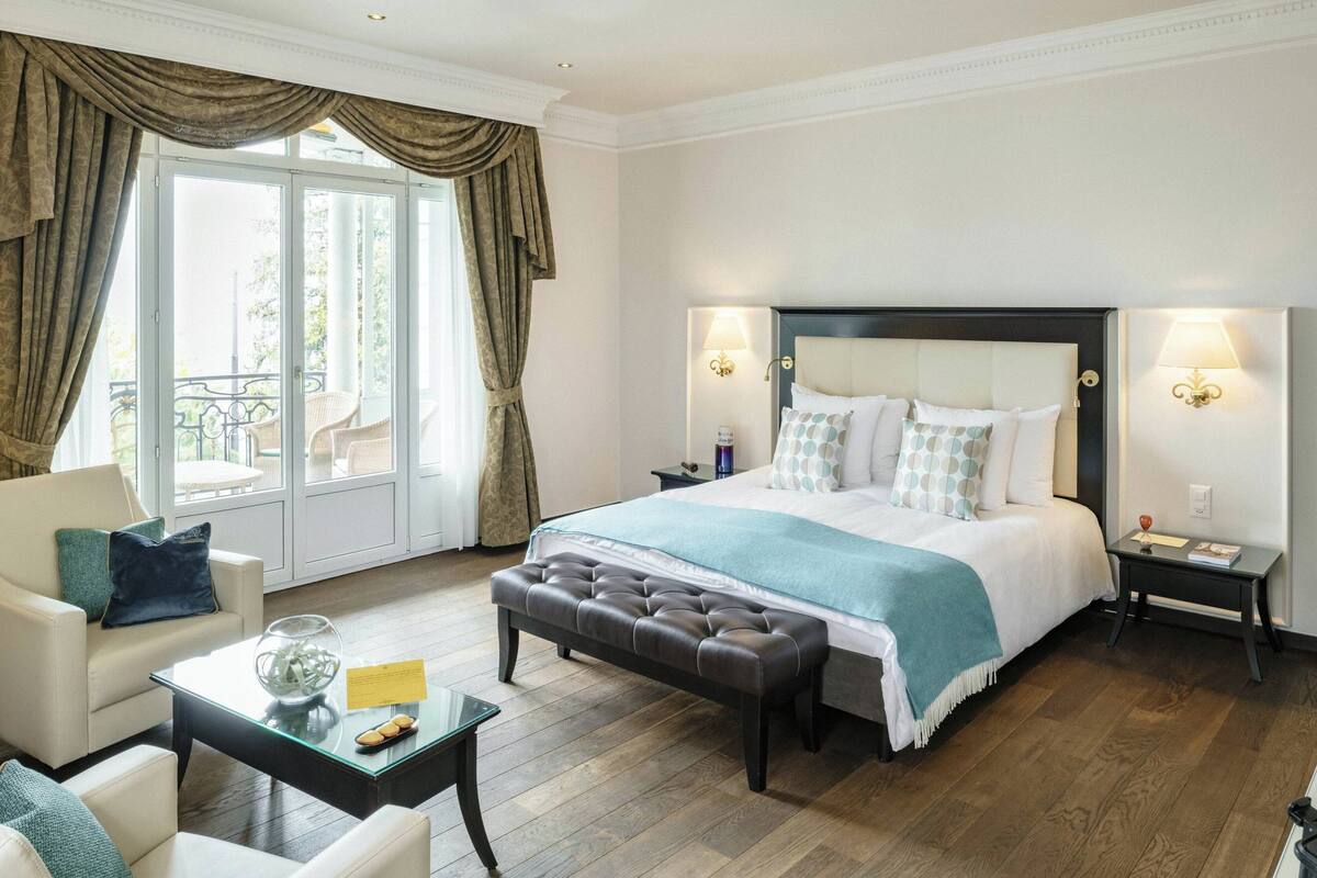 Habitación de hotel con una cama grande, dos mesitas de noche, un sofá, una mesa de centro y vistas al exterior a través de una puerta corrediza de vidrio.