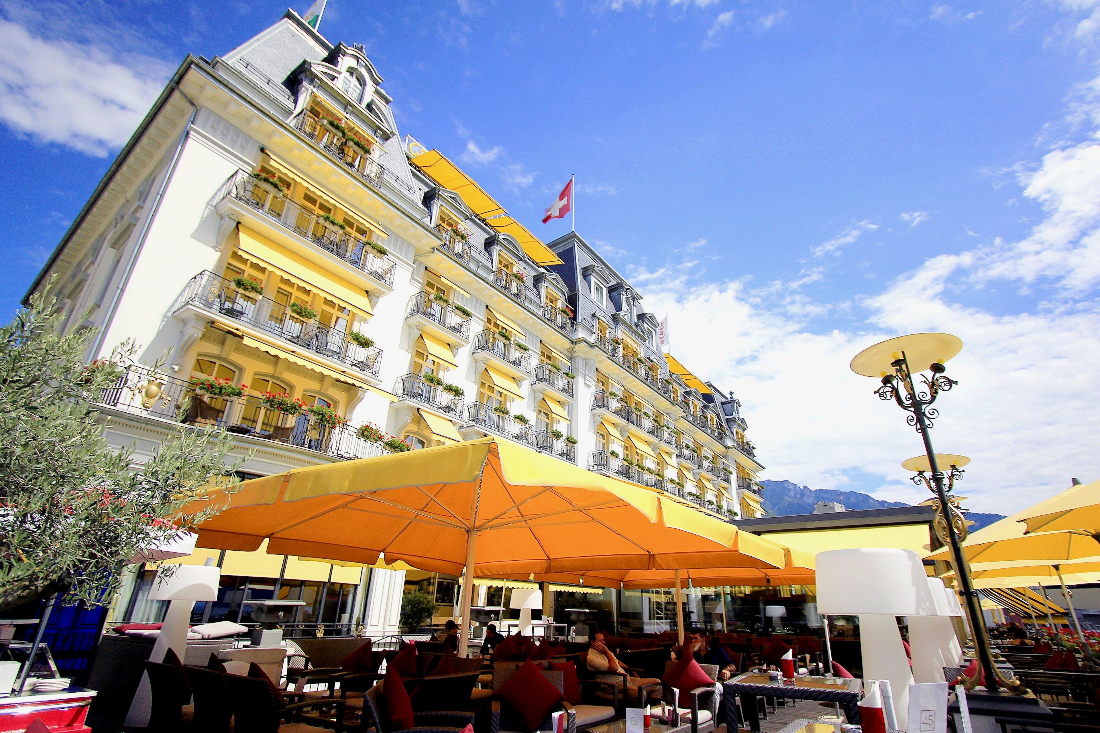 Ein Hotel mit Außenbereich, gelben Sonnenschirmen, Balkonen mit Pflanzen und einer Flagge.