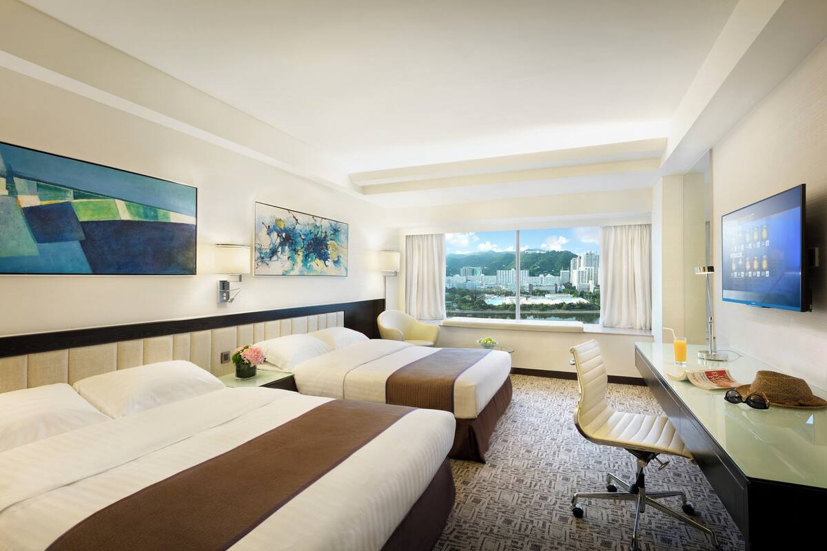 Habitación de hotel con dos camas, un escritorio, una silla, un televisor y una ventana con vistas a la ciudad.