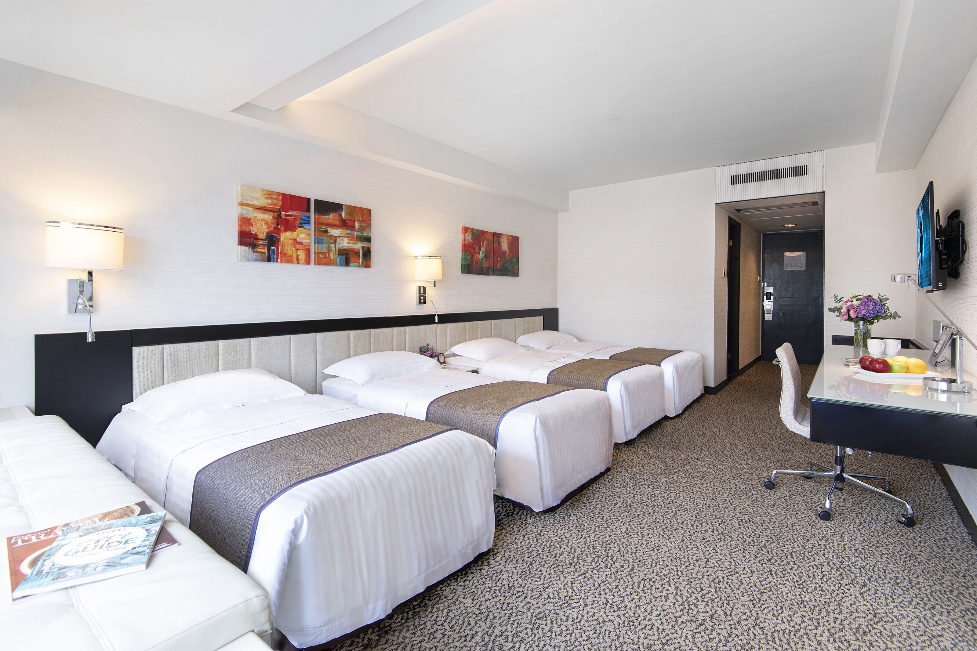 Una habitació d'hotel amb tres llits, una taula de treball amb una cadira, i obres d'art a les parets.