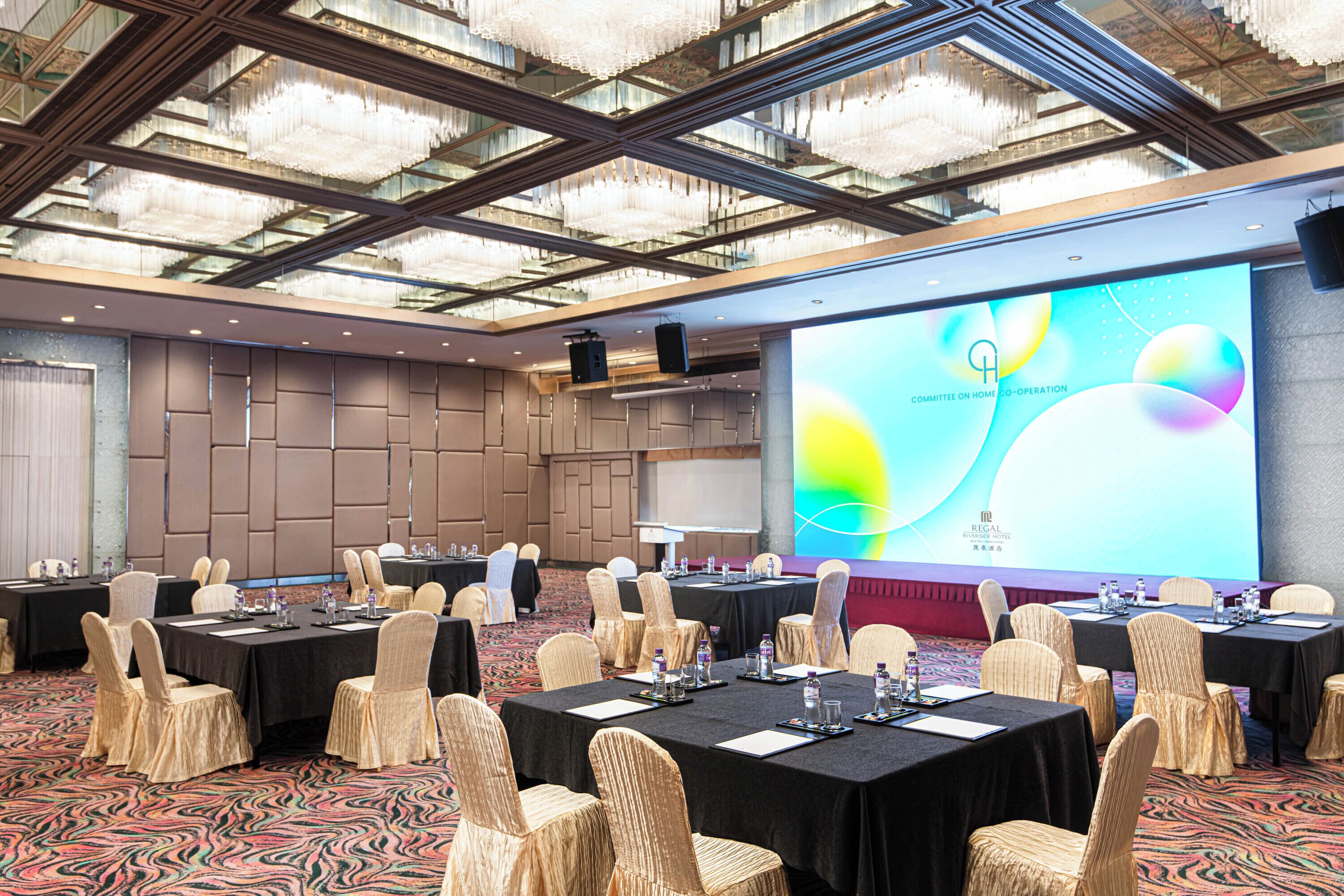 A conference room with tables, chairs, a large screen, and a chandelier.