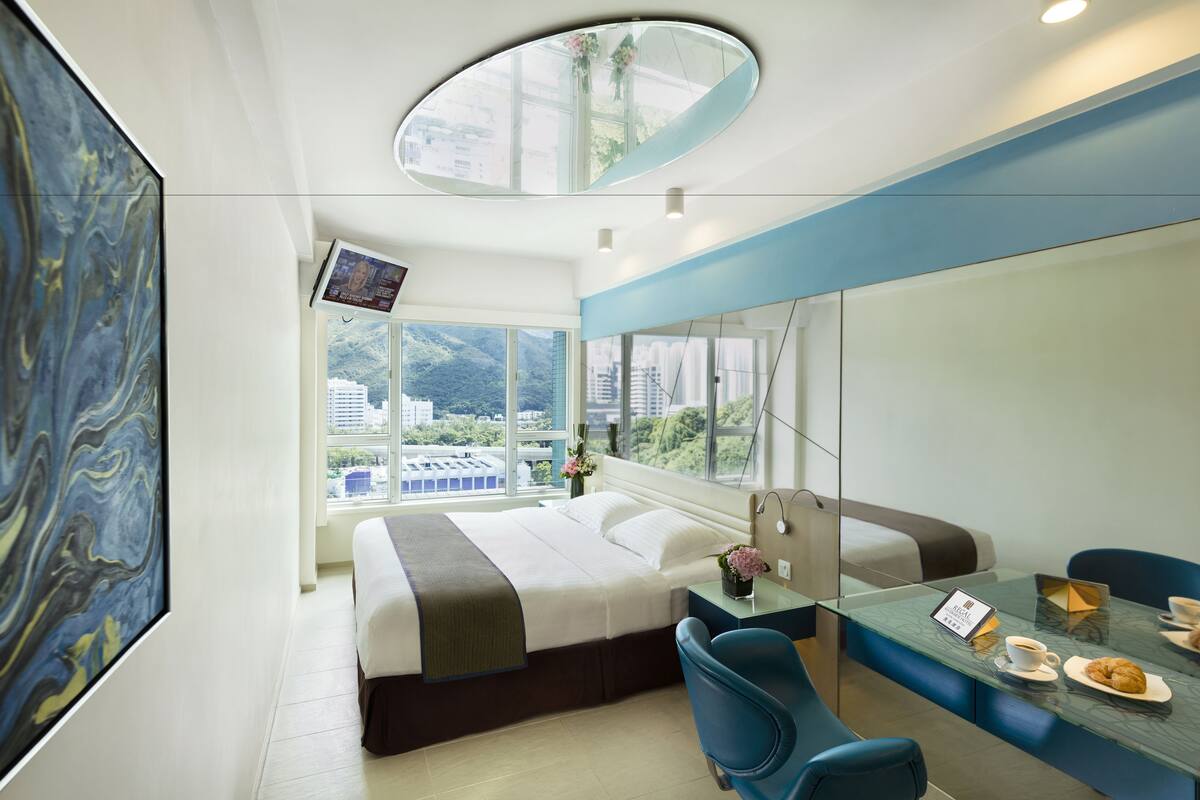 Habitación de hotel moderna con una cama grande, una mesa de vidrio con una taza de café y un croissant, un televisor en la pared y vistas de la ciudad a través de amplias ventanas.