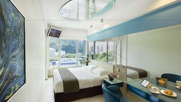 A modern hotel room with a large bed, a glass table with a coffee cup and croissant, a TV on the wall, and a view of the city through large windows.