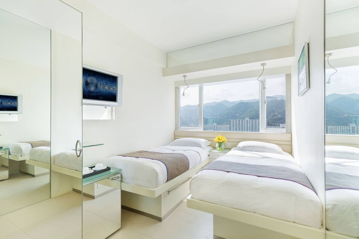 Una habitación de hotel moderna con dos camas, un ventanal con vistas a la ciudad y las montañas, un televisor en la pared y un espejo que refleja la habitación.
