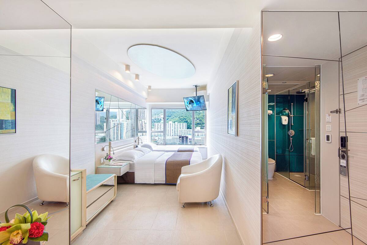 Una habitación de hotel moderna con una cama, dos sillones blancos, un escritorio y un amplio ventanal con vistas a la ciudad.