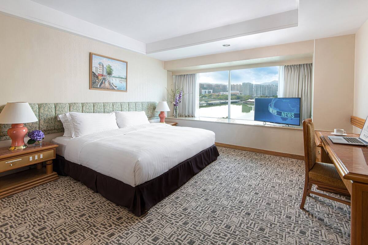 Habitación de hotel con una cama grande, un escritorio con una computadora portátil, un televisor y vistas a un paisaje urbano a través de un ventanal amplio.