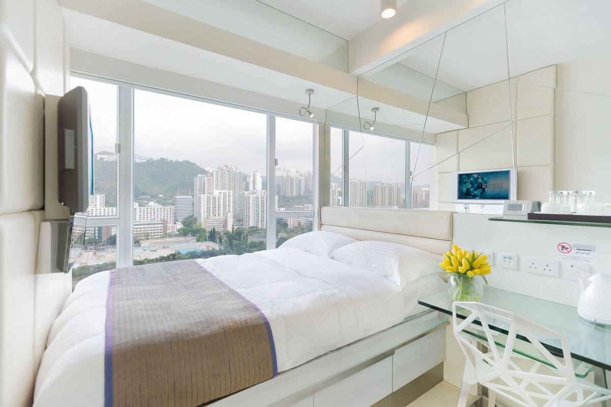 Habitación de hotel moderna con una cama grande, un escritorio de vidrio y vistas a la ciudad a través de amplios ventanales.