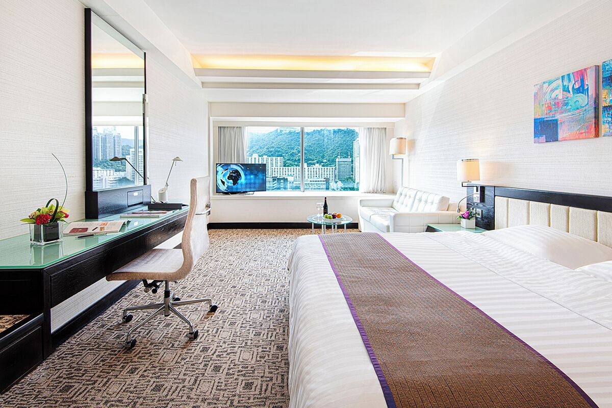 Habitación de hotel con una cama grande, un escritorio con computadora, una silla, un televisor y una ventana con vista a la ciudad.