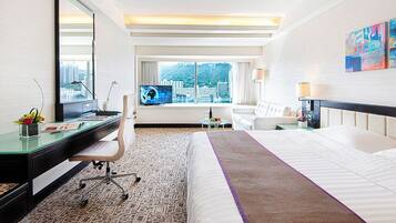 Habitación de hotel con una cama grande, un escritorio con computadora, una silla, un televisor y una ventana con vista a la ciudad.