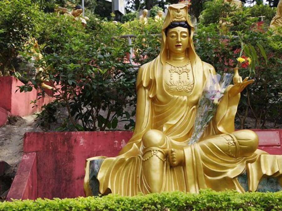 A large golden statue of a seated figure with a flower in hand, surrounded by greenery and other statues.