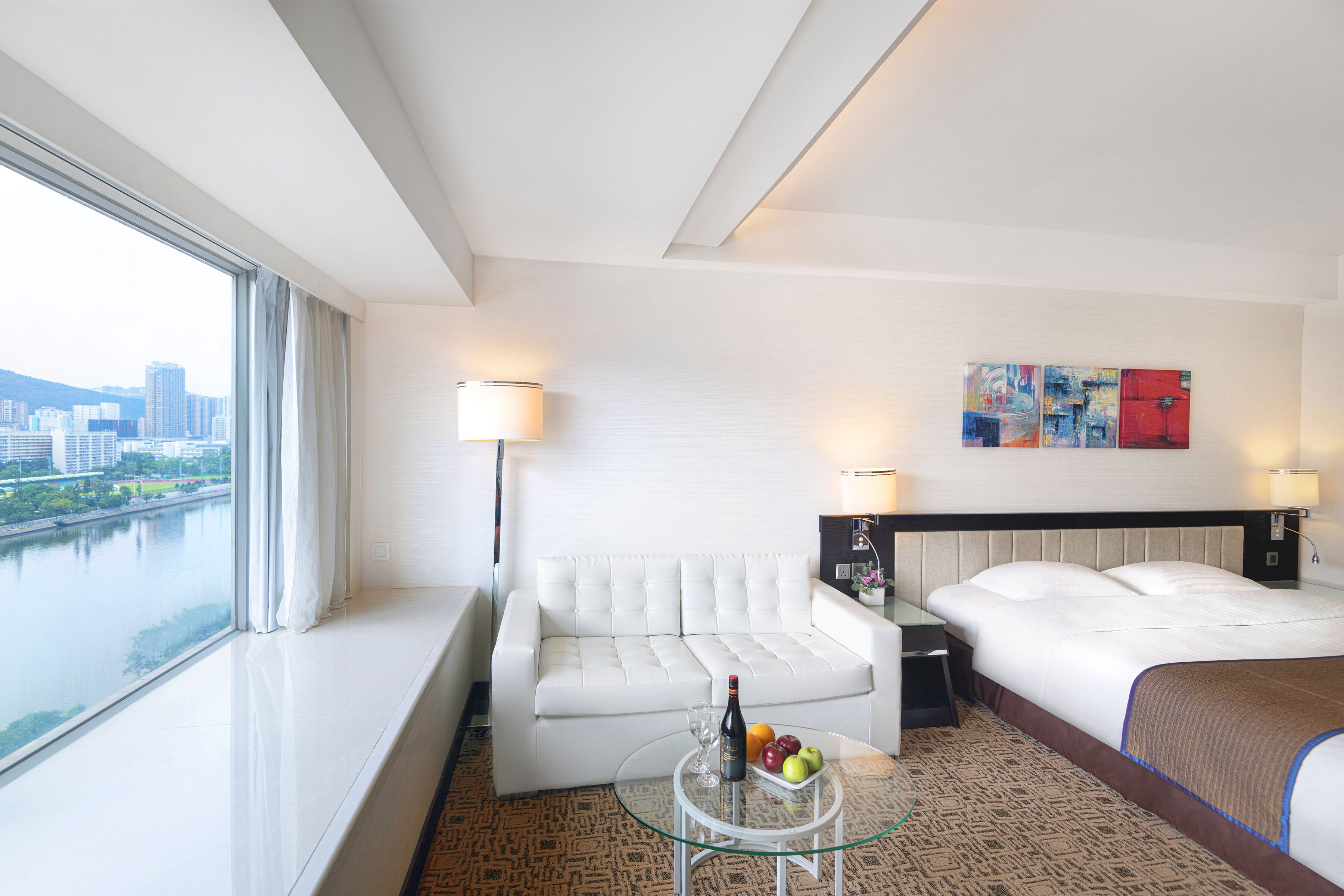A hotel room with a bed, a white sofa, a glass coffee table with fruits and a bottle, and a view of a cityscape through large windows.