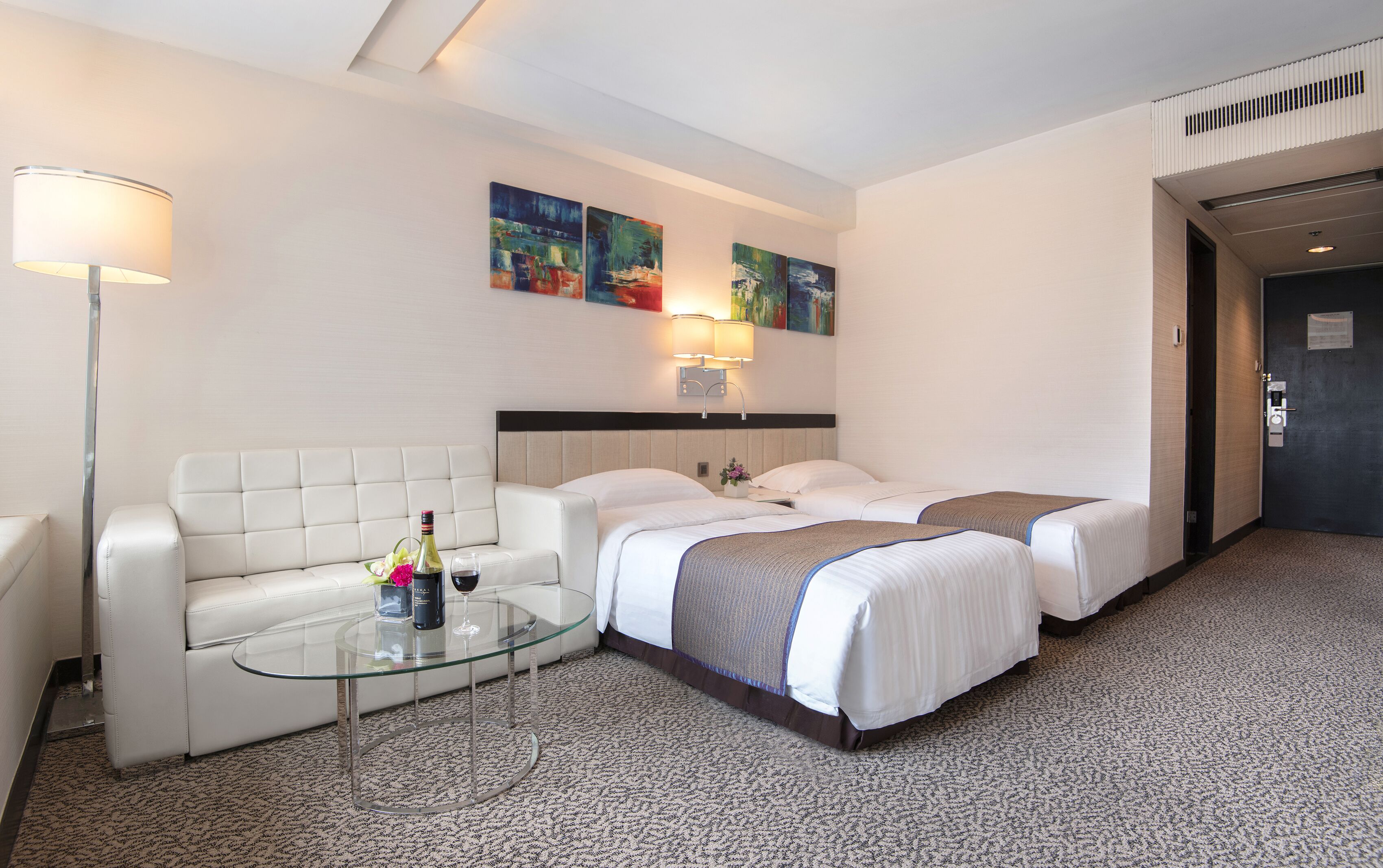 A hotel room with two beds, a glass coffee table with a bottle and glass, a white sofa, and artwork on the wall.