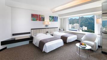 Habitación de hotel con dos camas, una mesita con fruta, una silla y un amplio ventanal con vista a la montaña.