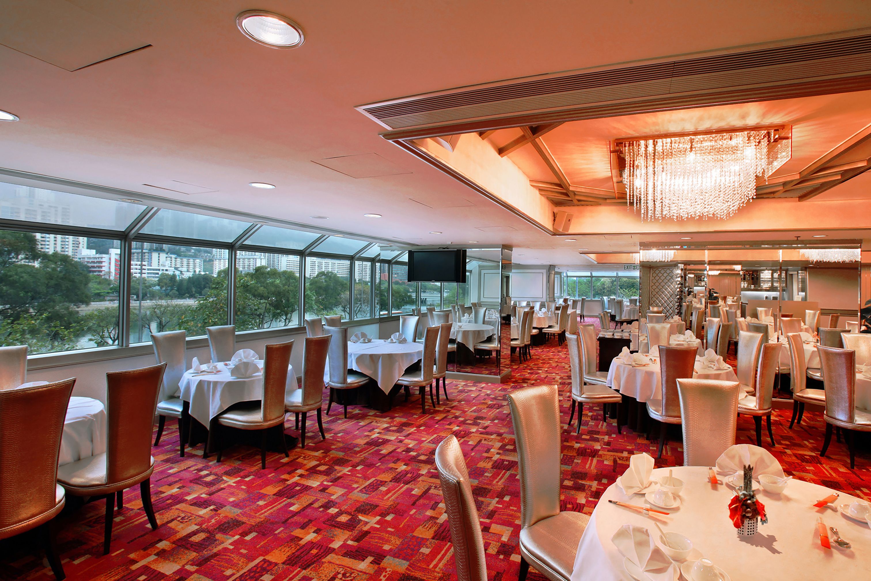 A spacious dining area with multiple tables and chairs, large windows offering a city view, and a modern chandelier.