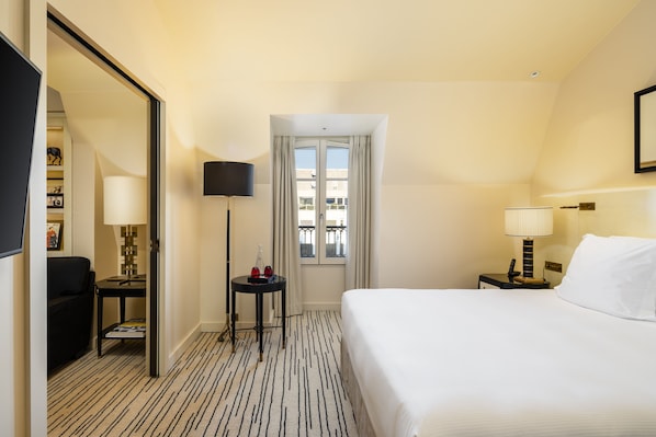 A hotel room with a bed, a desk, a chair, a TV, a window with curtains, and a door leading to another room. - Hôtel Montaigne (Paris)