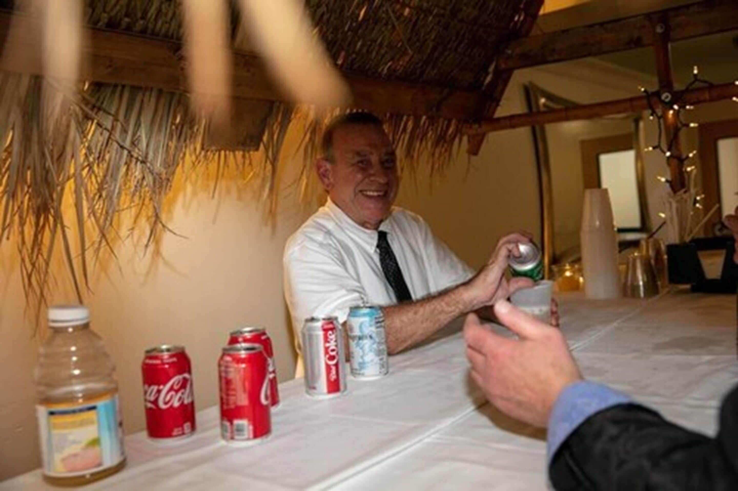 Una persona sentada a una mesa con una botella, una lata de Coca-Cola y una lata de Coca-Cola Light.