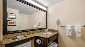 A hotel bathroom with a granite countertop sink, a large mirror, a towel rack, and a toilet paper dispenser.