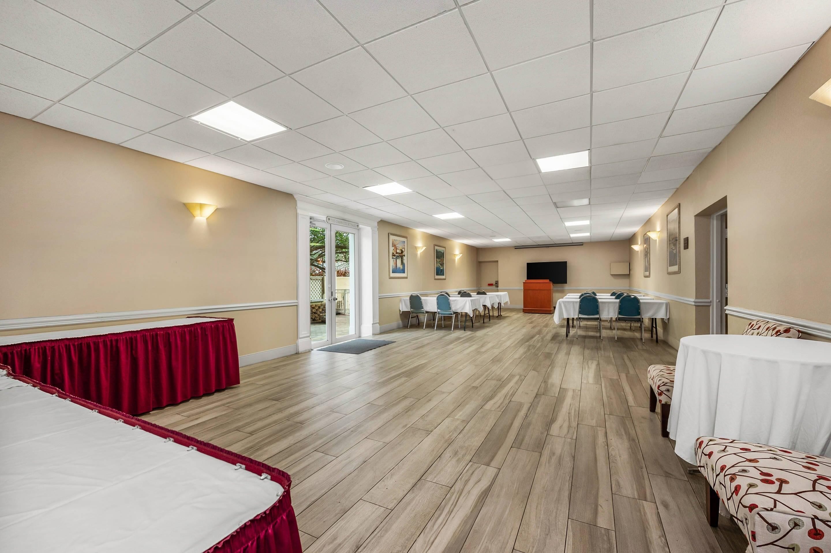 A spacious hotel room with a wooden floor, a red curtain, a white bed, and a dining area with tables and chairs.