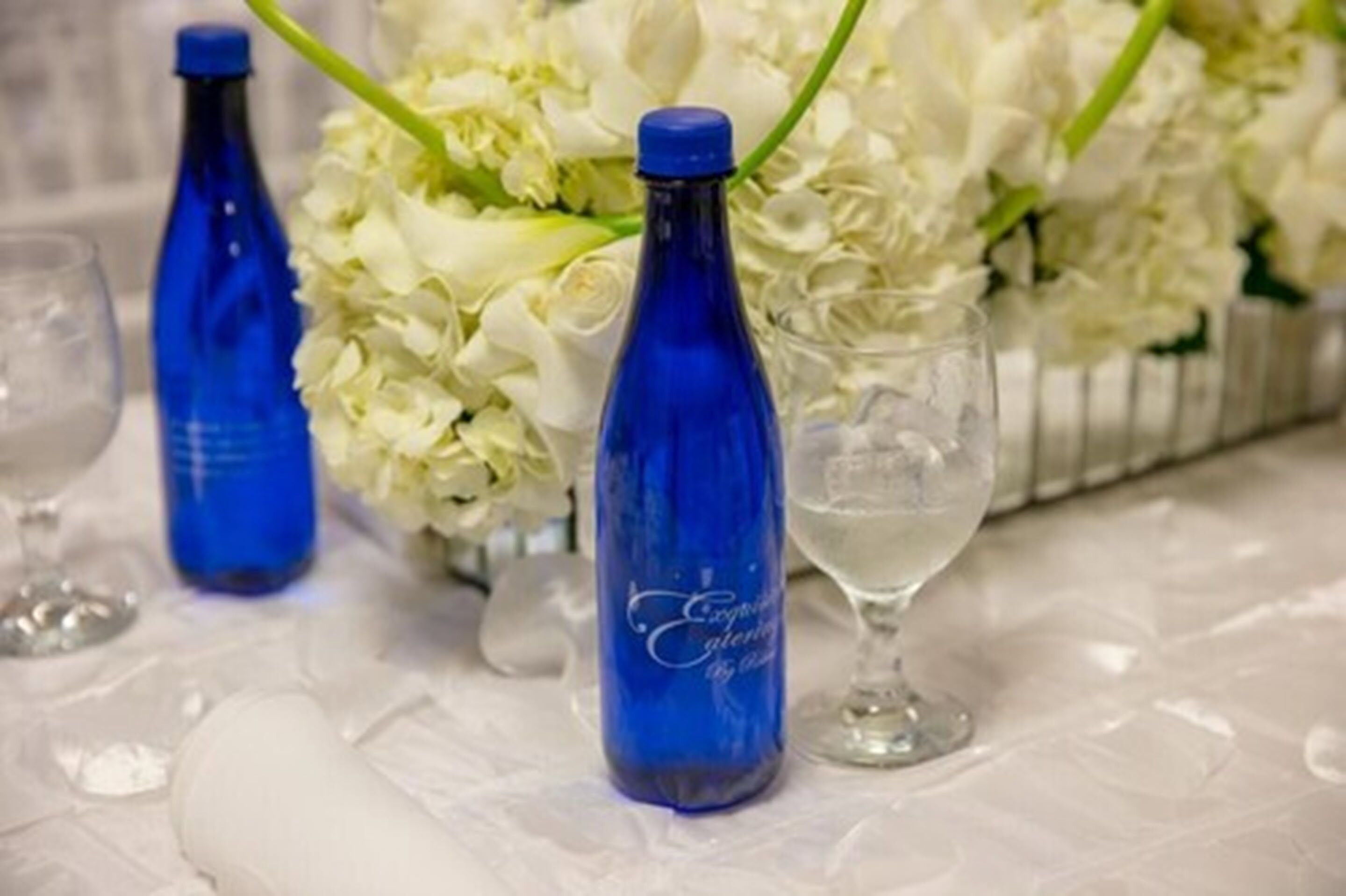 Une table avec des bouteilles bleues, un verre et un bouquet de fleurs blanches.