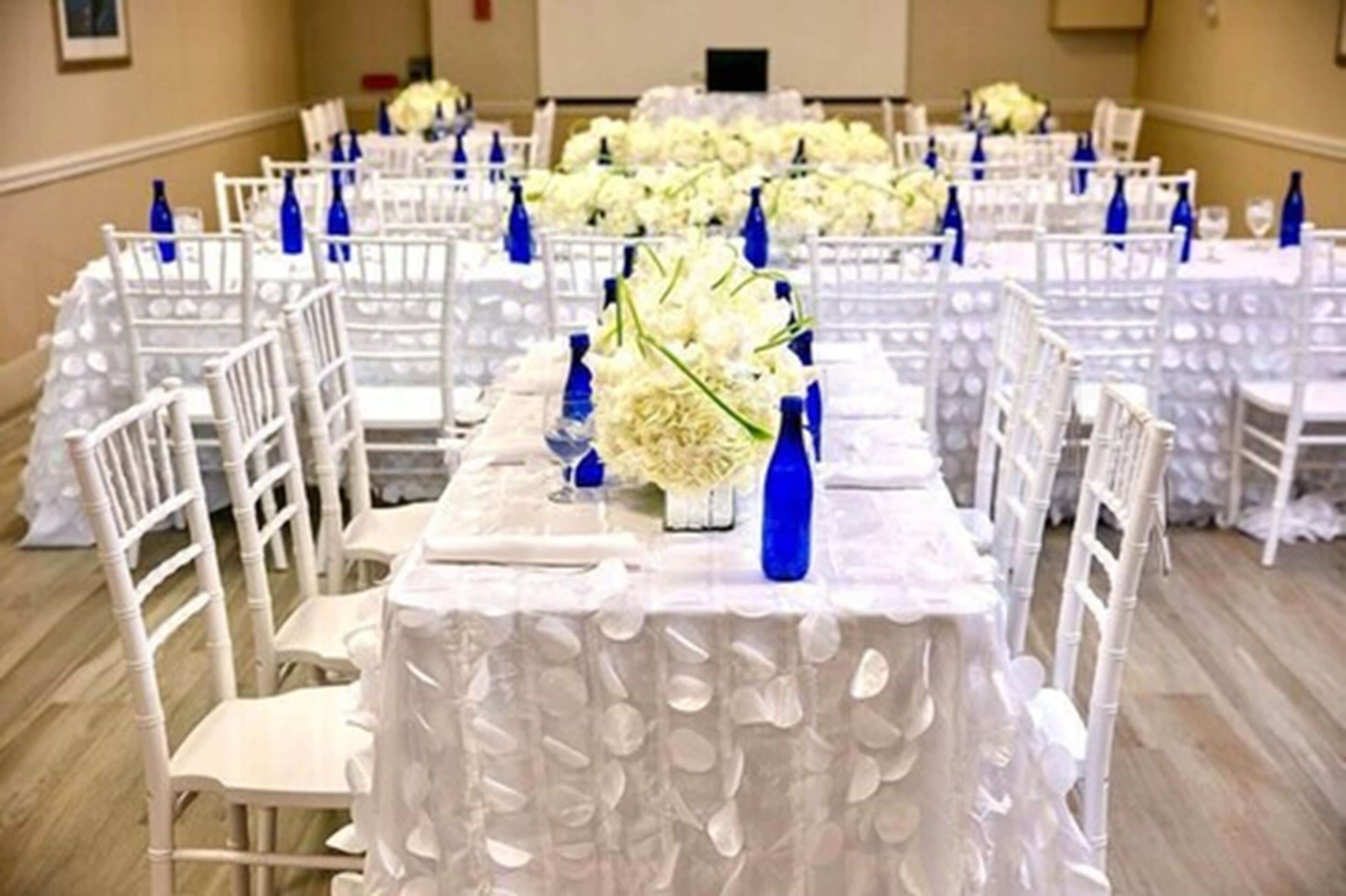 A banquet hall with white chairs and tables, each with a white tablecloth and blue bottle centerpieces.
