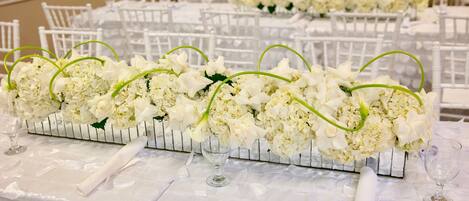 Une table de banquet dressée avec des nappes blanches, des chaises blanches et des centres de table floraux.