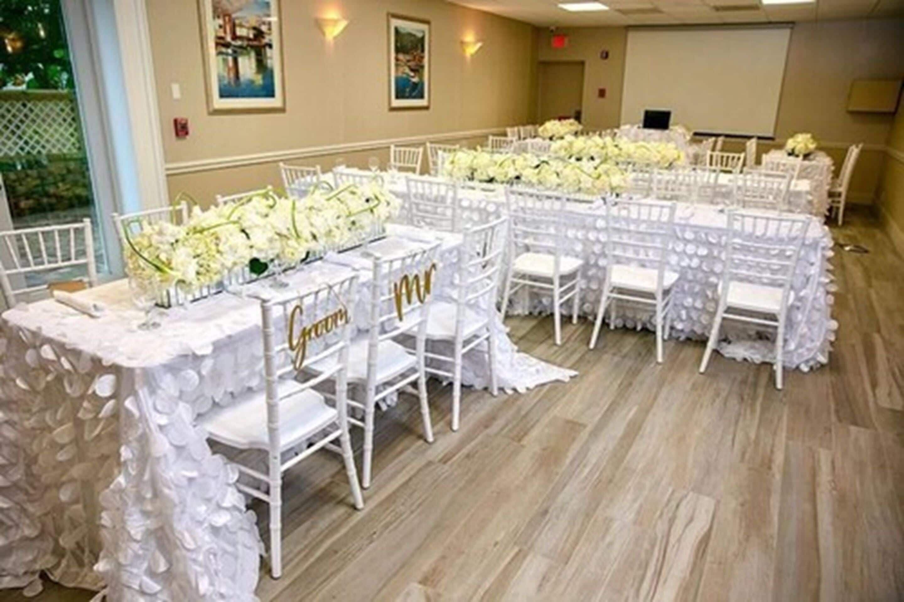 A banquet hall with white chairs and tables, a projector screen, and a large window with a view of greenery.