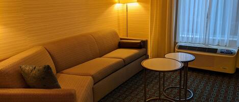 A hotel room with a sofa, a small table, a lamp, and an air conditioning unit.