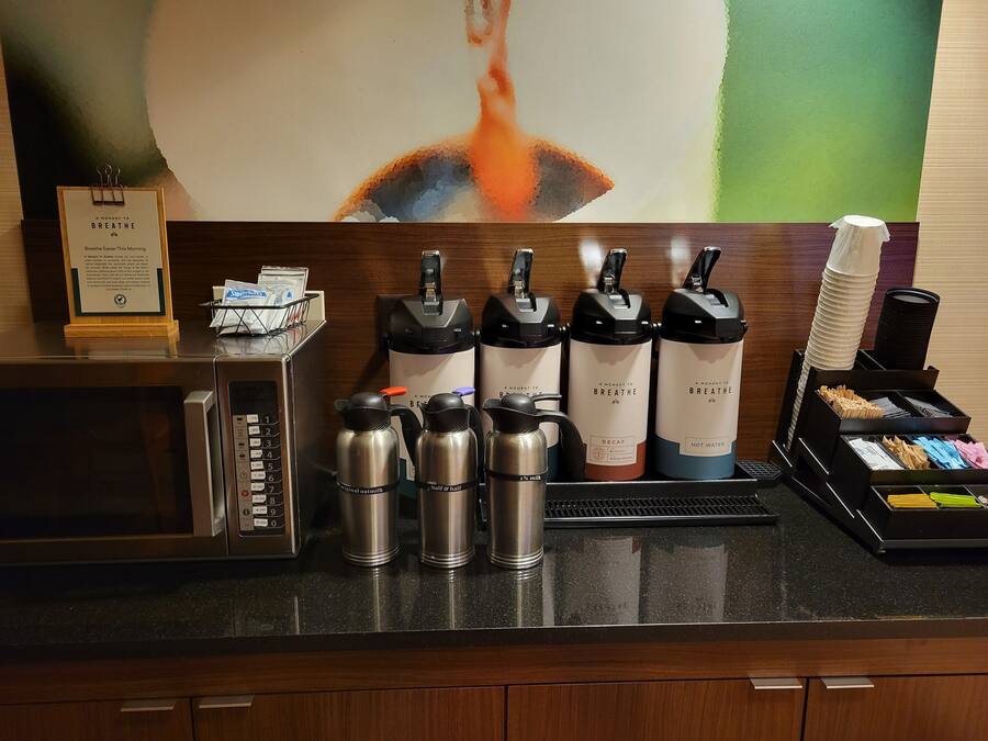Zona de recepción de hotel con estación de café que incluye termos de café descafeinado y agua caliente con la marca, un microondas y una pizarra con el menú.