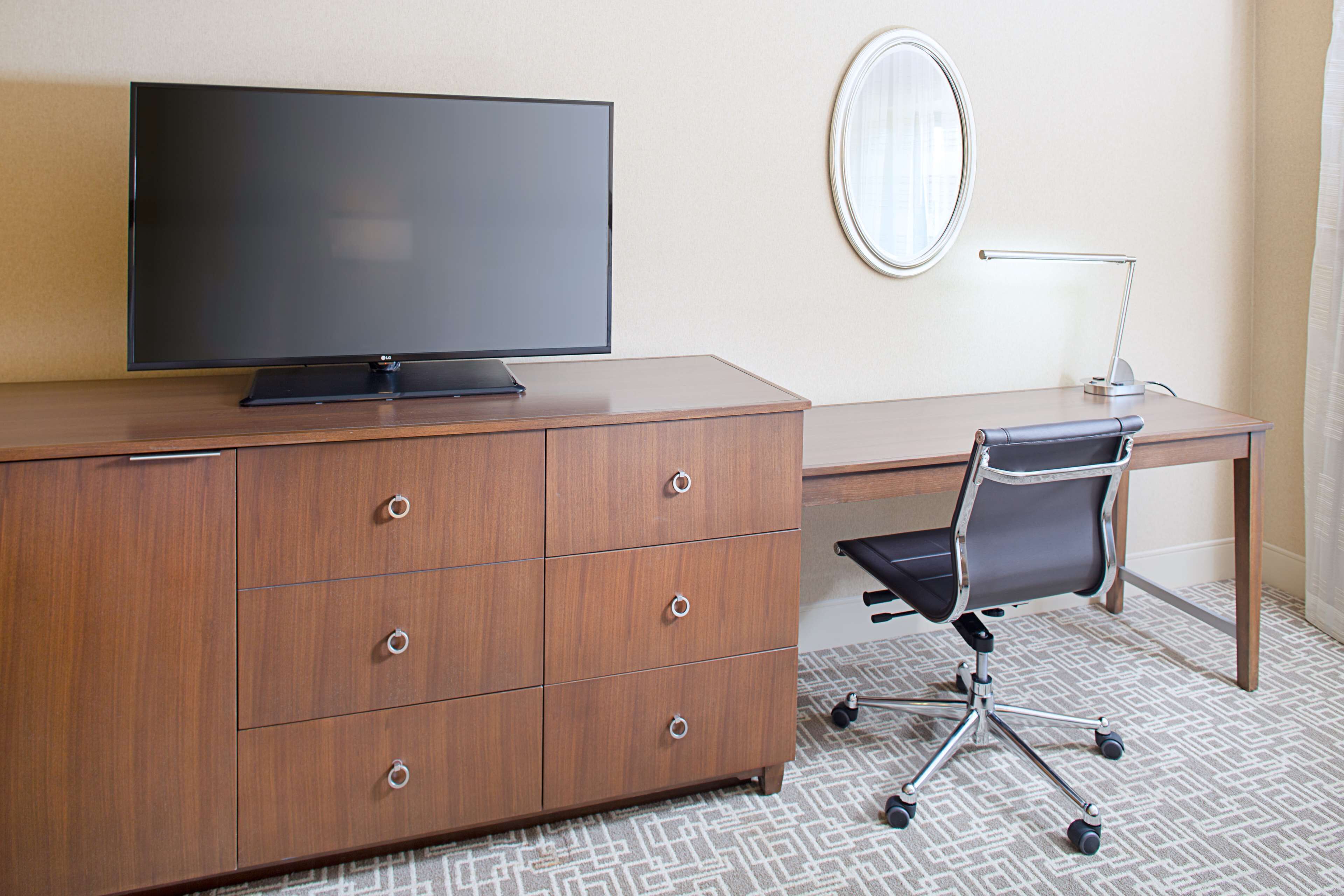A hotel room with a wooden desk, a flat-screen TV, a black office chair, a round mirror, and a desk lamp.
