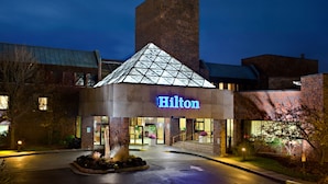 A Hilton hotel with a prominent glass pyramid entrance, a brick facade, and a well-lit exterior. - Hilton Boston/Dedham (Dedham)