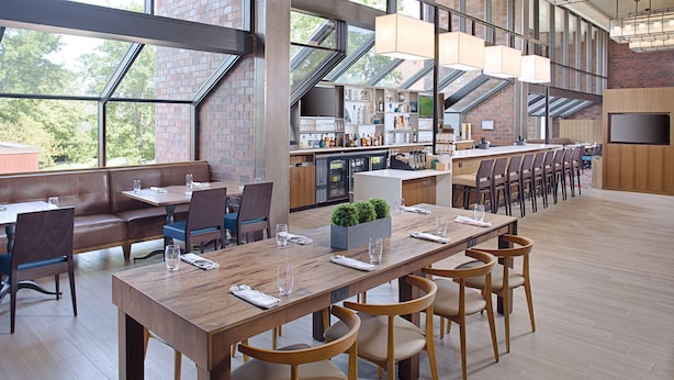 A spacious restaurant with high ceilings, large windows, and a long wooden table with chairs. - Hilton Boston/Dedham (Dedham)