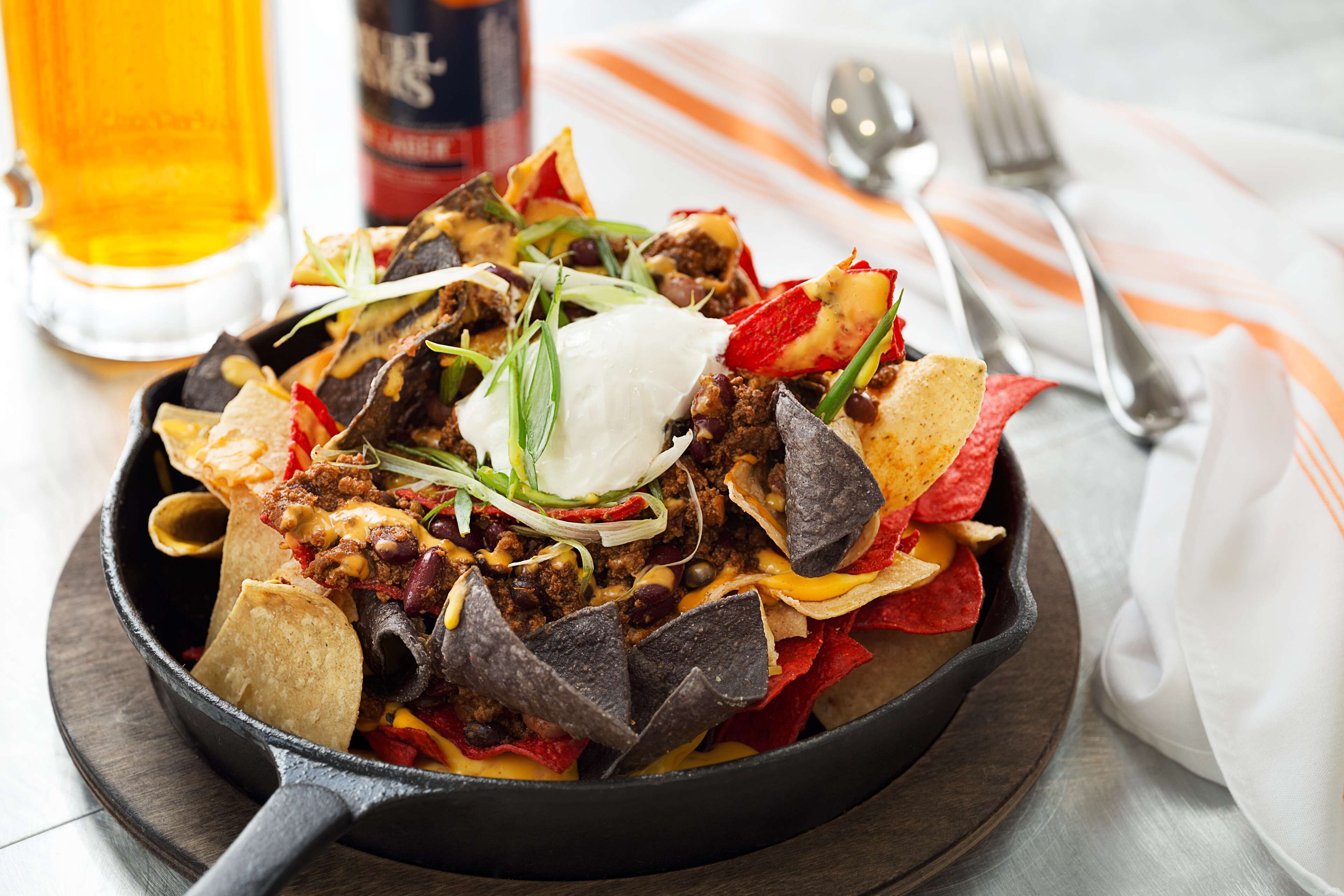 Una sartén con nachos cubiertos de queso, frijoles y carne, acompañada de una cerveza y una botella de salsa.