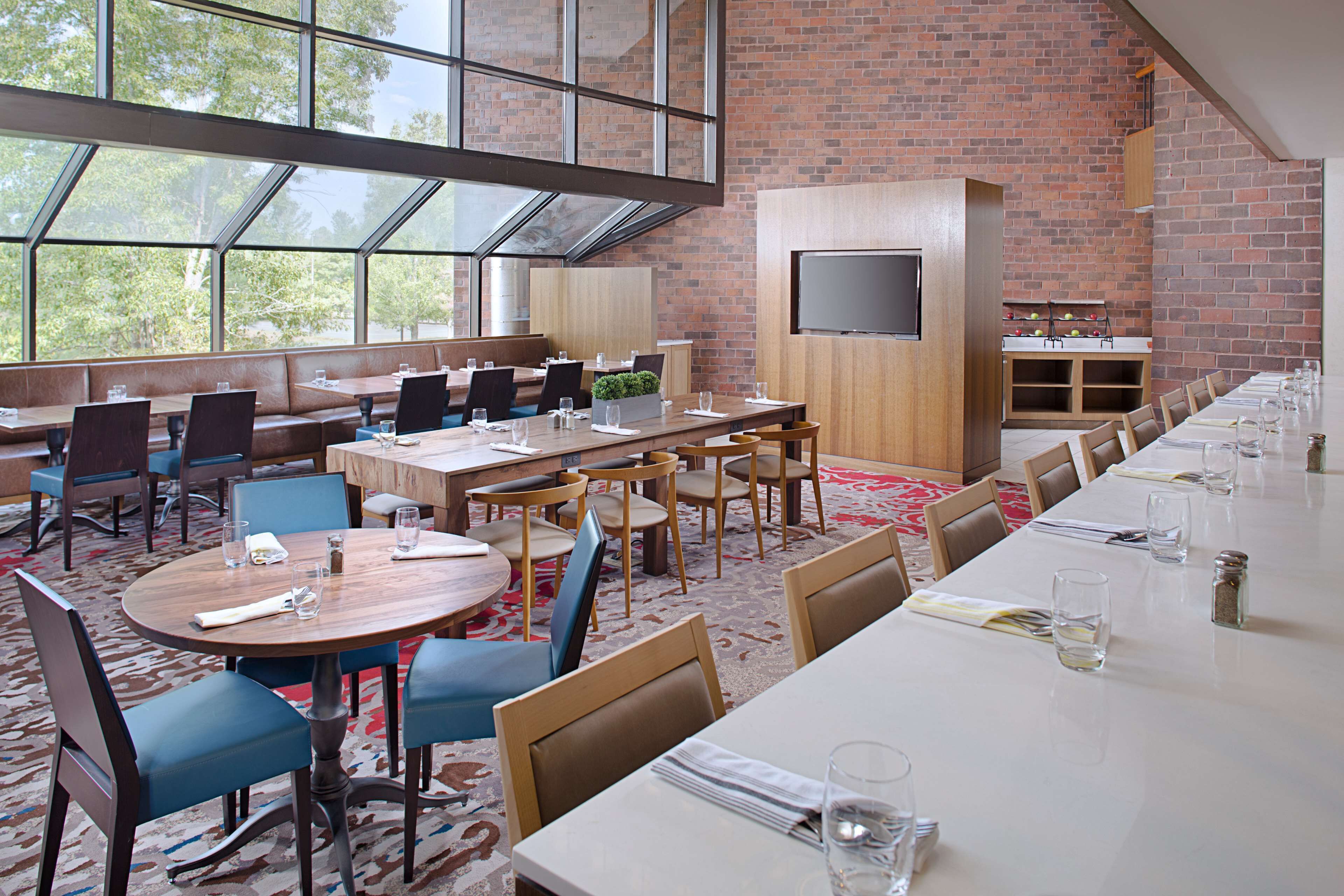 A spacious dining area with high ceilings, large windows, and a brick wall. Tables are set with chairs, and a flat-screen TV is mounted on the wall.