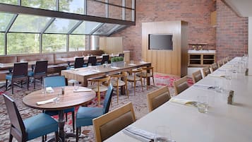 A spacious dining area with high ceilings, large windows, and a brick wall. Tables are set with chairs, and a flat-screen TV is mounted on the wall.