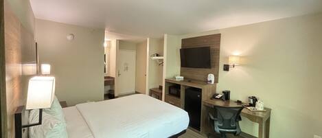 A hotel room with a bed, desk, chair, TV, and a small kitchenette.