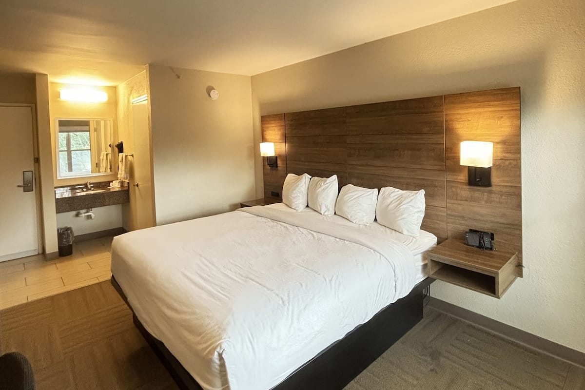 Une chambre d’hôtel avec un grand lit, une tête de lit en bois, une table de chevet, un miroir et une salle de bain équipée d’un lavabo et d’un porte-serviettes.