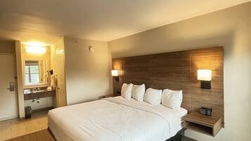 Habitación de hotel con una cama grande, cabecera de madera, mesita de noche, espejo y baño con lavabo y toallero.