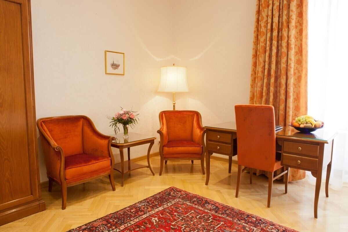 Una habitación con un escritorio de madera, dos sillas, una lámpara, un florero con flores y una alfombra estampada.