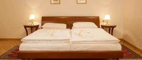 A double bed with white linens and two bedside lamps, framed pictures on the wall, and a patterned rug on the floor.