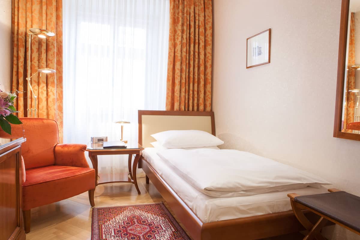 A hotel room with a single bed, a wooden nightstand, a desk with a chair, a desk lamp, and a large window with curtains.