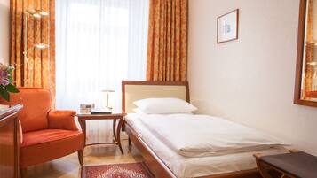 A hotel room with a single bed, a wooden nightstand, a desk with a chair, a desk lamp, and a large window with curtains.