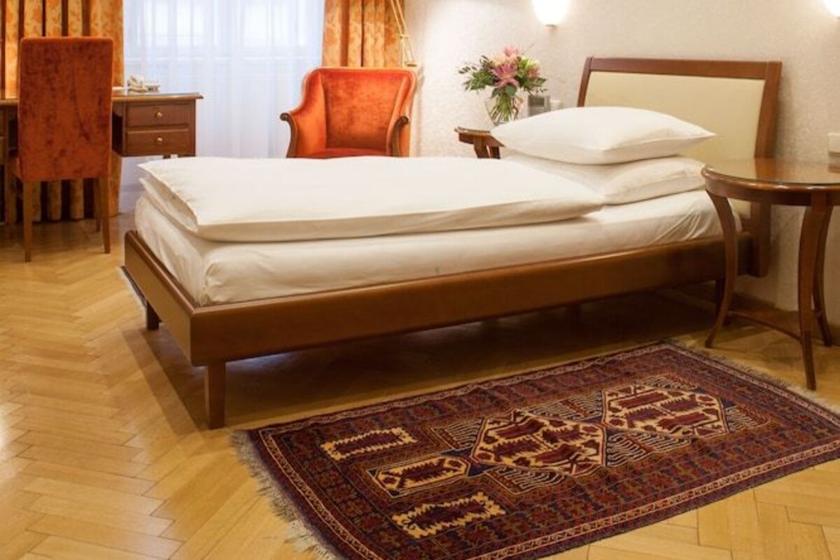Habitación de hotel con cama, escritorio, silla, mesita de noche, lámpara y alfombra.
