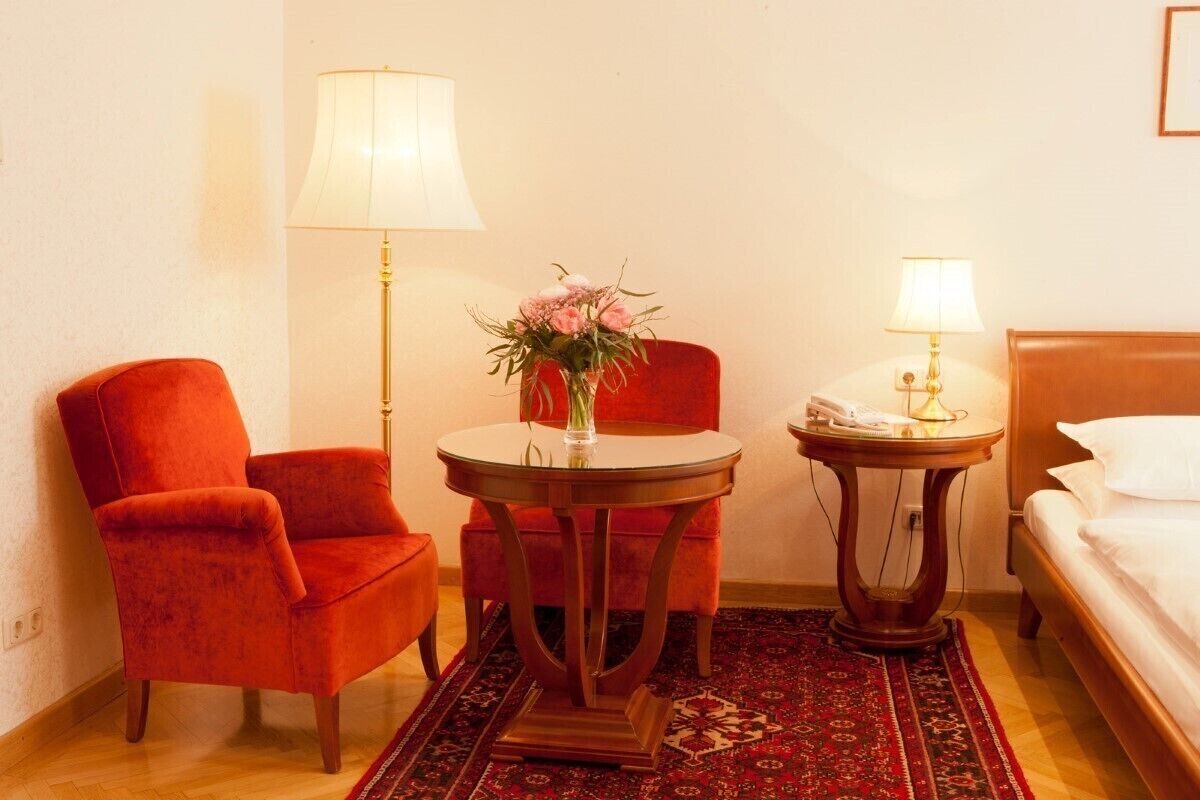 Quarto de hotel com uma poltrona vermelha, uma mesa redonda de madeira com um vaso de flores, uma mesa redonda pequena com um abajur e uma cama com roupa de cama branca.