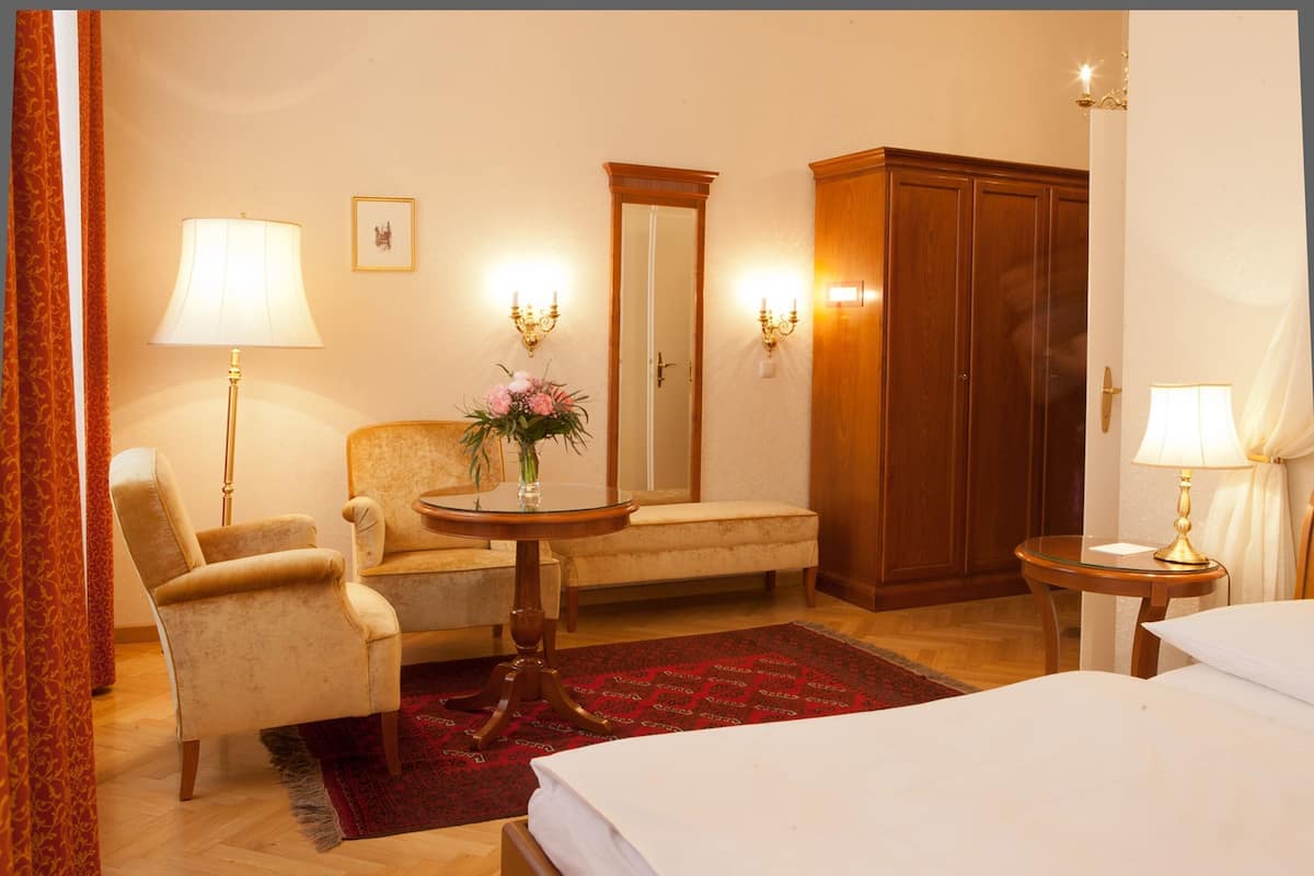 A hotel room with a bed, a chair, a small round table with a vase, a wooden wardrobe, and a mirror.