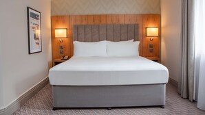 A hotel room with a large bed, wooden headboard, two bedside lamps, a framed picture on the wall, and a patterned carpet.