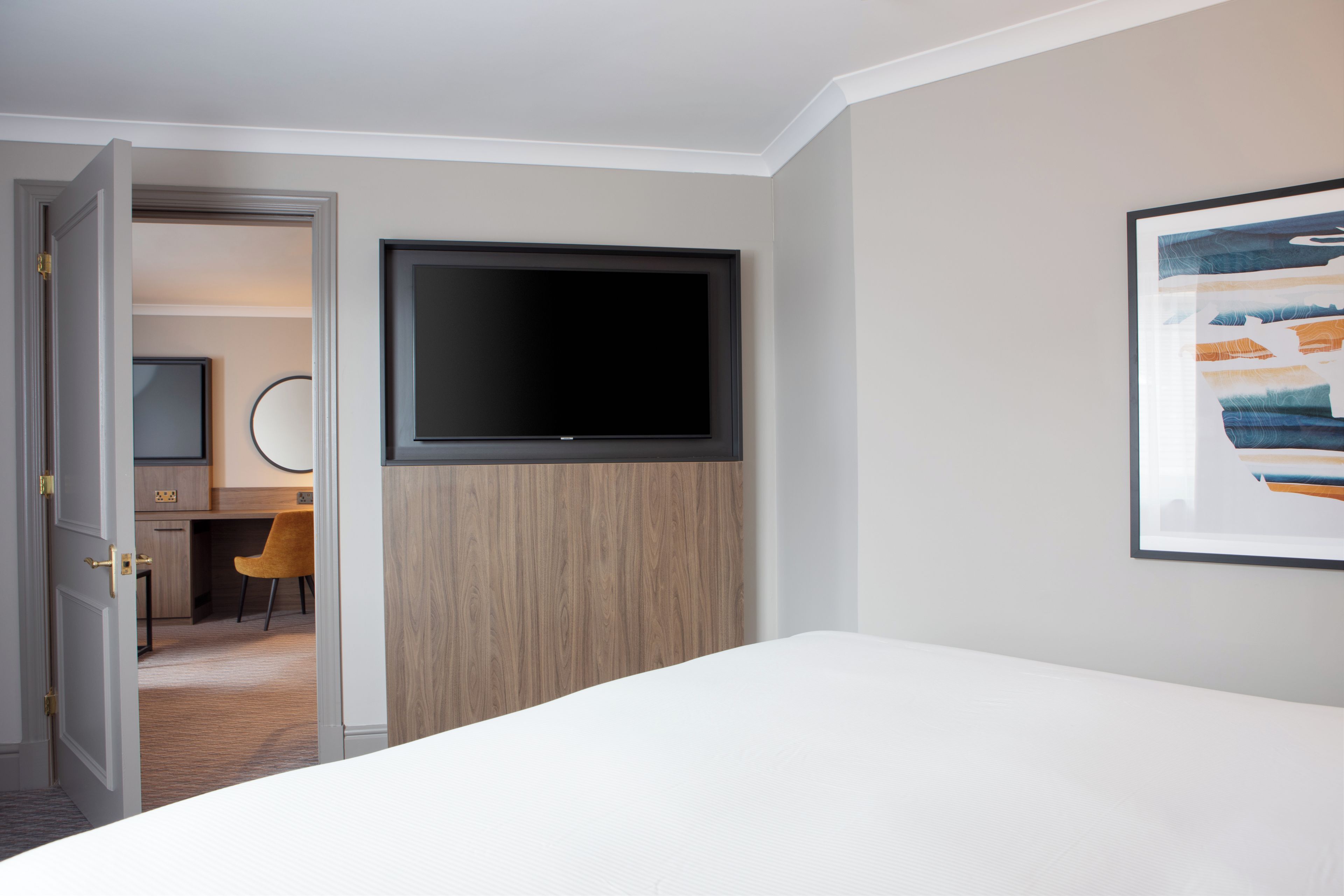 Une chambre d’hôtel avec un lit, un téléviseur fixé sur un panneau en bois, un bureau avec une chaise et un tableau encadré au mur.