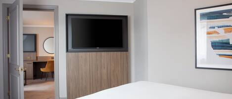 A hotel room with a bed, a TV mounted on a wooden panel, a desk with a chair, and a framed picture on the wall.