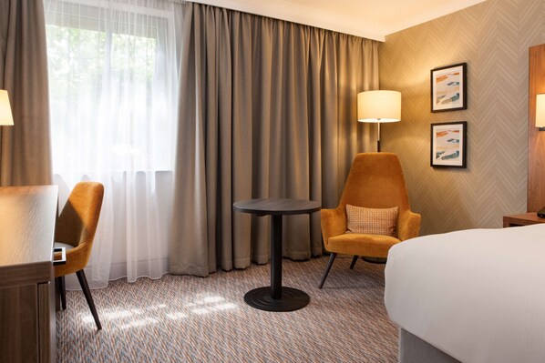 A hotel room with a bed, a round table, two chairs, a lamp, and a view of the outdoors through sheer curtains.