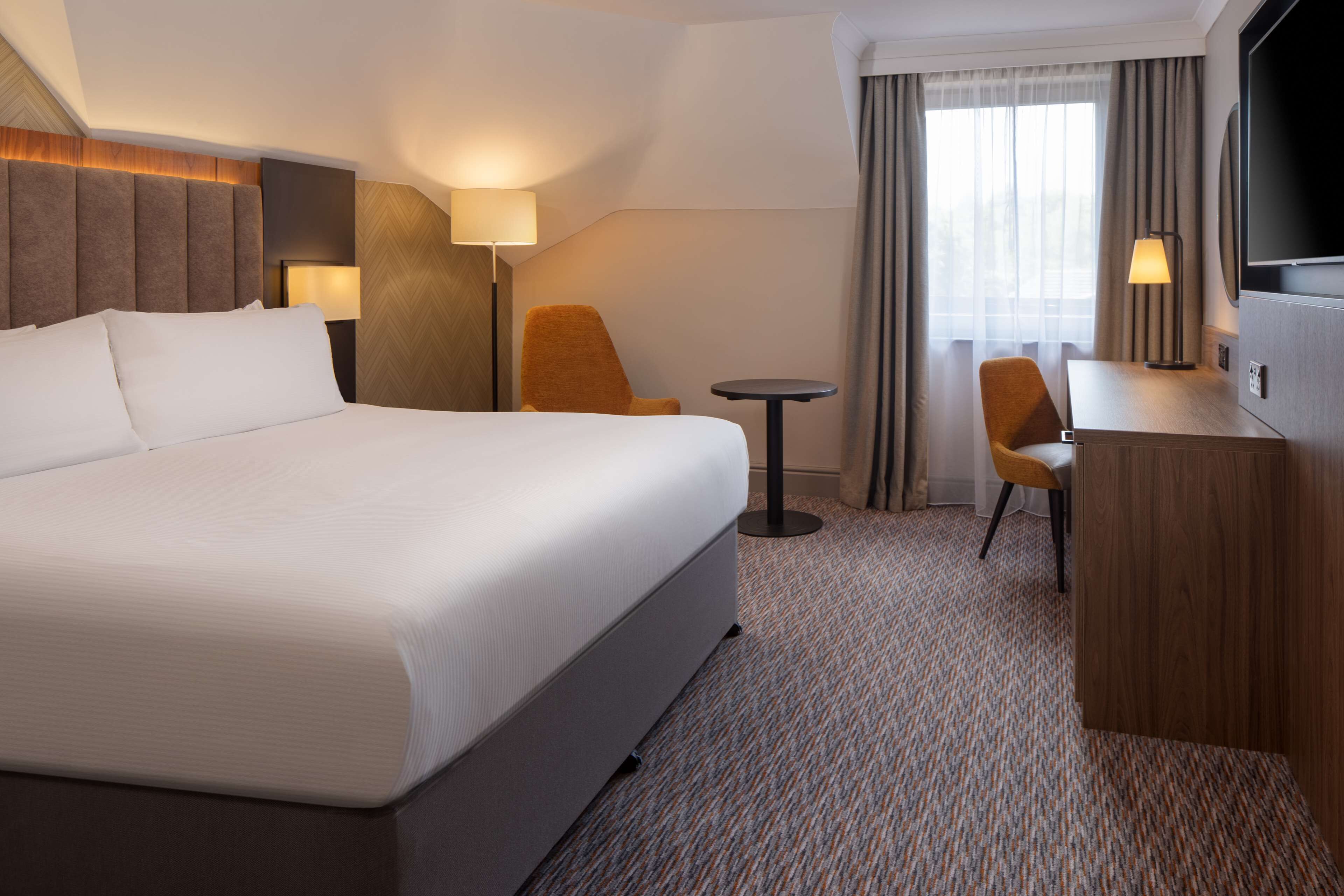 A hotel room with a large bed, two chairs, a small table, a TV, and a lamp.